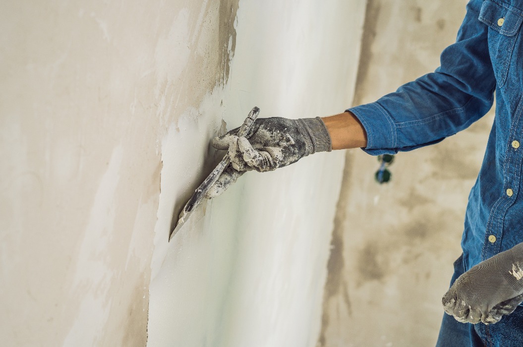 Meister trägt weiße Spachtelmasse auf eine Wand auf und verstreicht sie mit einem Spachtel in einem Raum eines renovierten Hauses bei Tag